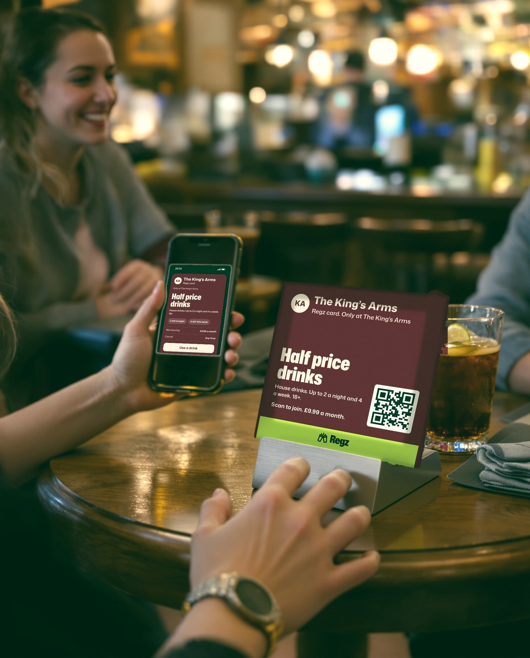 A customer holding up a phone showing The King's Arms Regz card, beside a table stand with its QR code